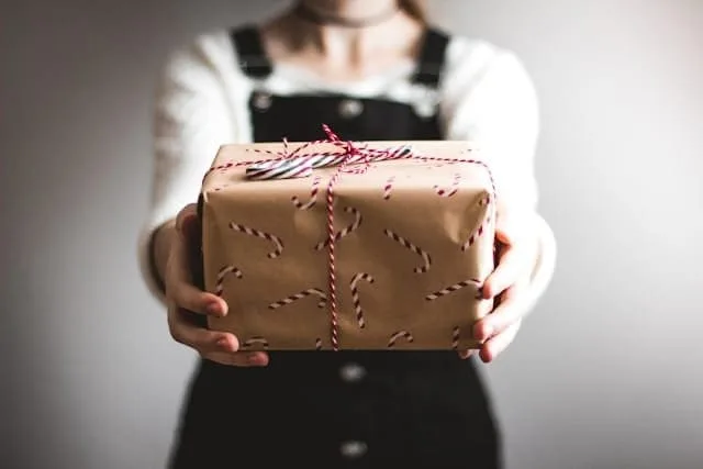 Uma pessoa segurando uma caixa de presentes, como se fosse entregá-la a quem está na sua frente. A imagem sugere a ideia de como usar possessivos em inglês.